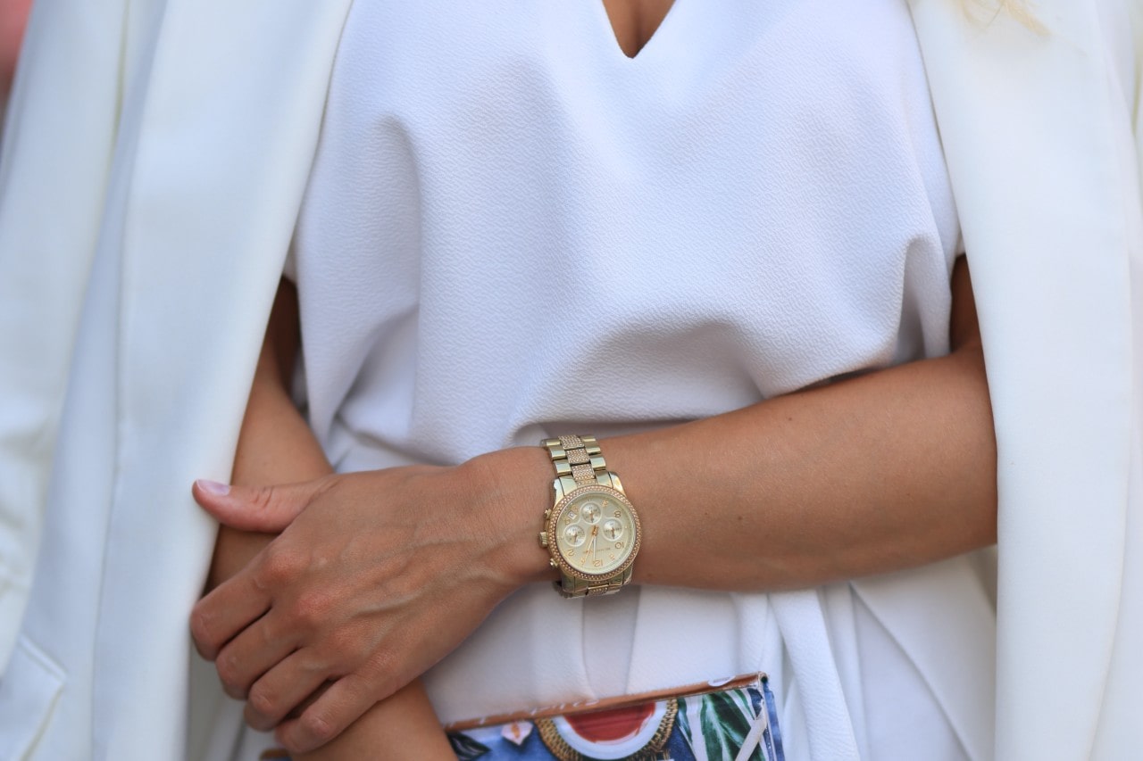 Close up of a woman wearing white and with a gold watch on her wrist and holding a colorful clutch in other hand