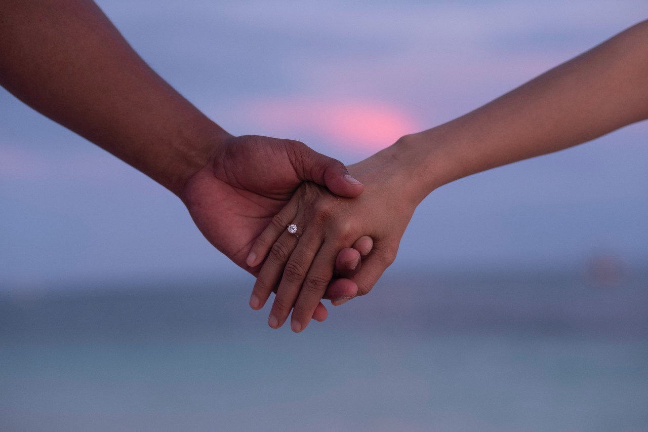 Couple holding hands in front of a beautiful sunset. Shown is a brilliant diamond
