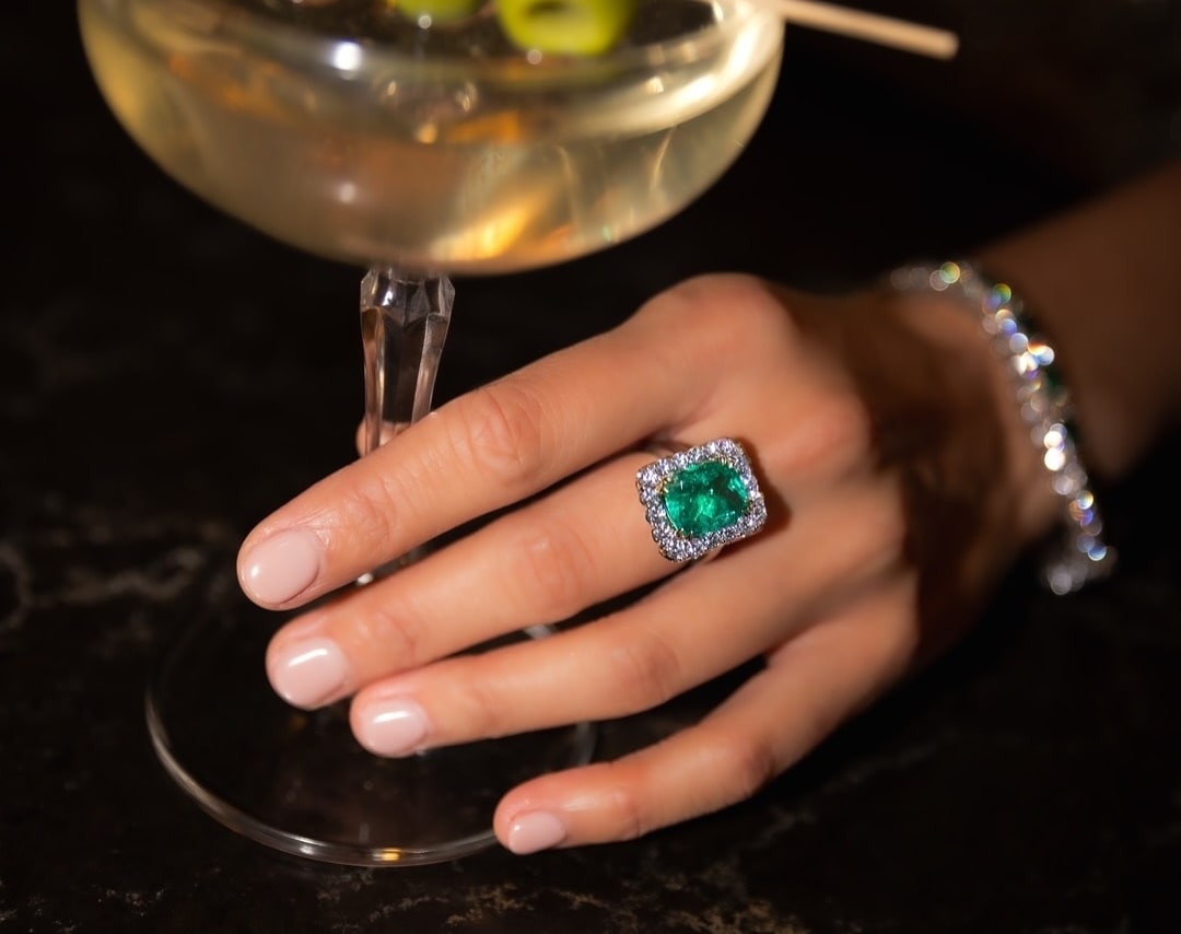 A woman holding onto a martini with an oval cut emerald ring with a diamond halo and a diamond bracelet as well