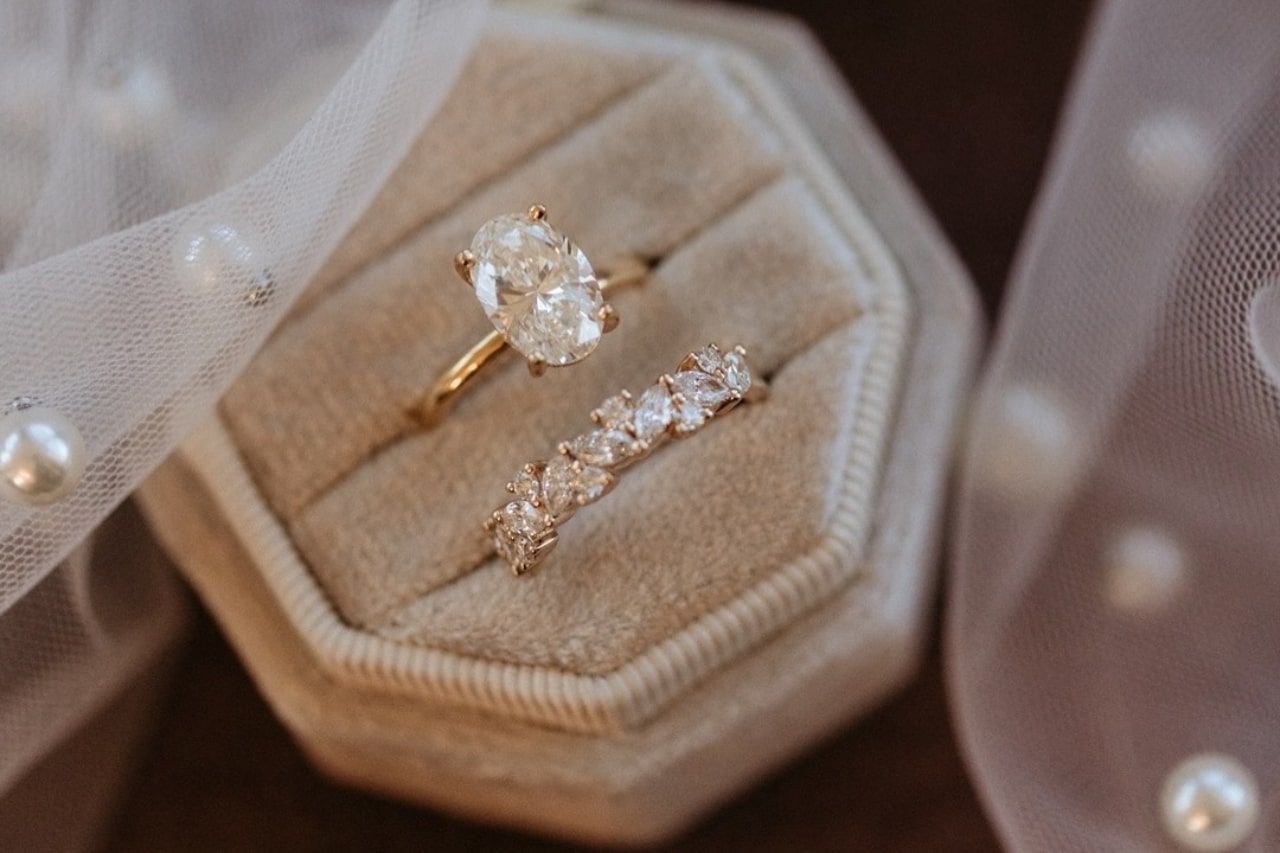 A close-up of an elegant oval engagement ring and accompanying wedding band displayed in-box beneath a veil adorned with pearls.